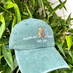 May include: A green baseball cap with an embroidered image of Winnie the Pooh holding a honey pot. The text "mama bear est 2024" is embroidered on the front of the cap.