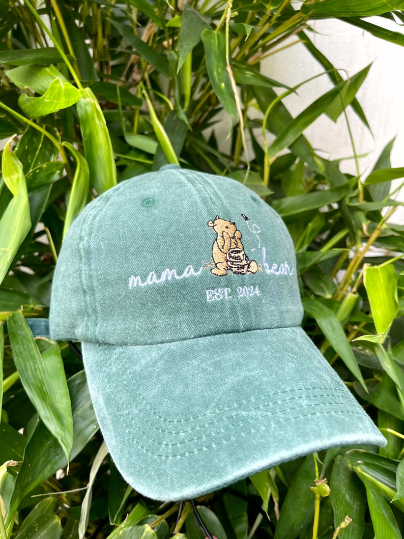 May include: A green baseball cap with an embroidered image of Winnie the Pooh holding a honey pot. The text "mama bear est 2024" is embroidered on the front of the cap.