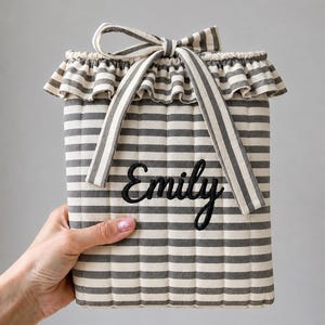 May include: A rectangular quilted fabric pouch featuring a grey and white horizontal striped pattern. The top edge has a ruffled trim and a matching fabric tie. The name Emily is embroidered in black cursive script on the front of the pouch.