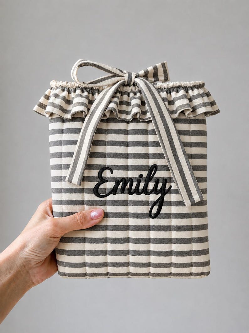 May include: A rectangular quilted fabric pouch featuring a grey and white horizontal striped pattern. The top edge has a ruffled trim and a matching fabric tie. The name Emily is embroidered in black cursive script on the front of the pouch.
