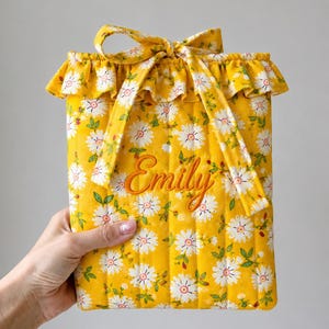 May include: A quilted yellow fabric pouch featuring a white daisy floral pattern with green leaves. The pouch has a ruffled top edge, a matching fabric tie closure, and the name Emily embroidered in orange cursive script on the front center.