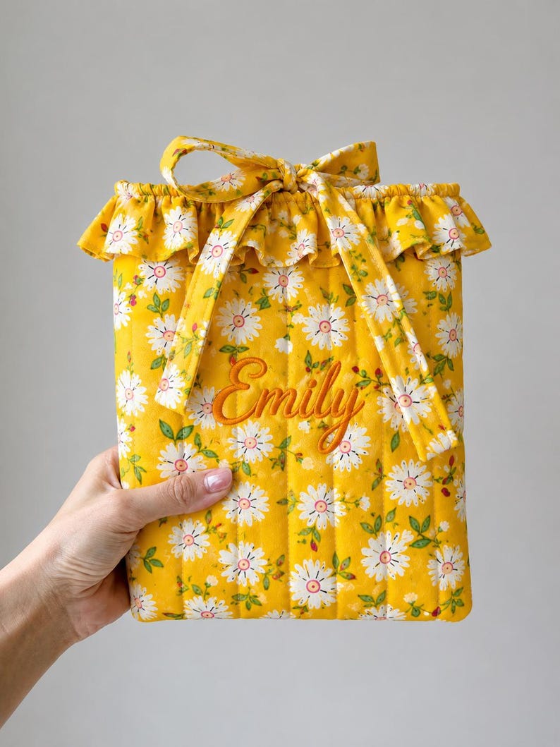 May include: A quilted yellow fabric pouch featuring a white daisy floral pattern with green leaves. The pouch has a ruffled top edge, a matching fabric tie closure, and the name Emily embroidered in orange cursive script on the front center.