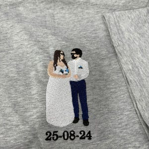 May include: A heather gray t-shirt featuring an embroidered design of two figures standing side by side. One figure wears a long white dress and the other wears a white shirt with dark blue pants. The date 25-08-24 is embroidered in black below.