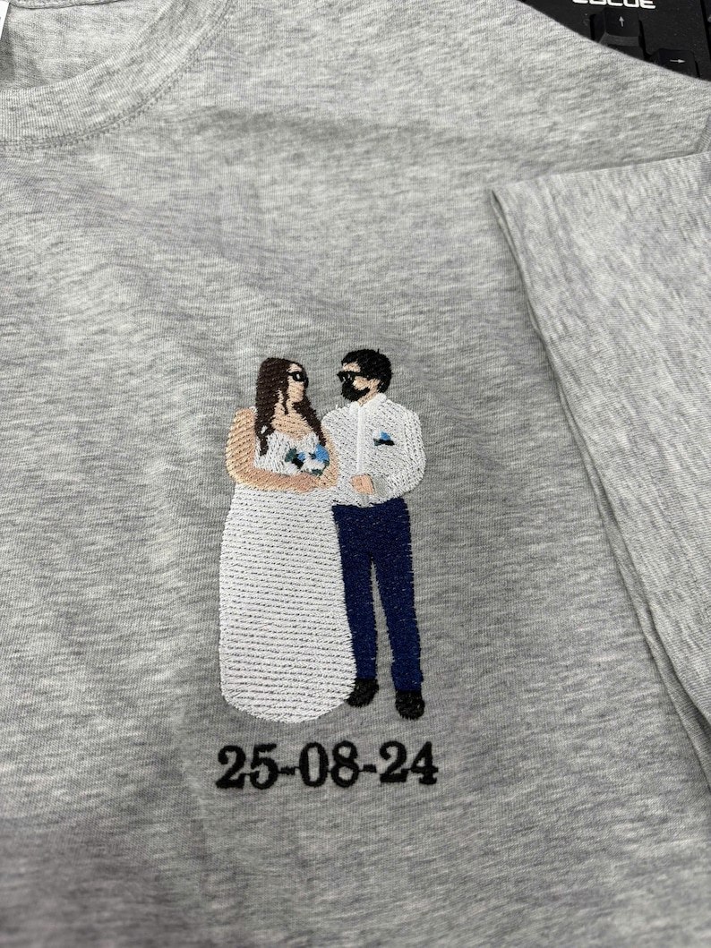 May include: A heather gray t-shirt featuring an embroidered design of two figures standing side by side. One figure wears a long white dress and the other wears a white shirt with dark blue pants. The date 25-08-24 is embroidered in black below.
