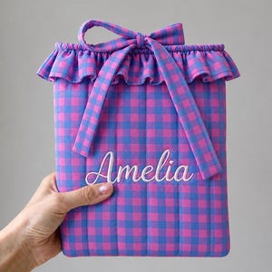 May include: A rectangular quilted pouch featuring a pink and blue gingham pattern. The top edge has a ruffled trim and a fabric tie closure. The name Amelia is embroidered in white cursive script across the center of the front panel.