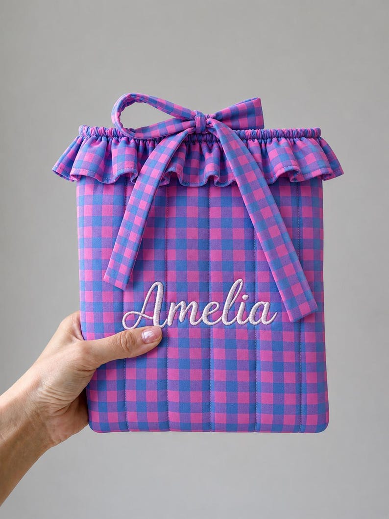 May include: A rectangular quilted pouch featuring a pink and blue gingham pattern. The top edge has a ruffled trim and a fabric tie closure. The name Amelia is embroidered in white cursive script across the center of the front panel.
