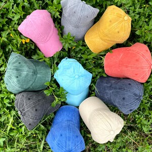 May include: Ten different colored baseball caps arranged in a circle on a bed of green grass. The caps are made of a soft, textured fabric and come in a variety of colors, including blue, green, red, pink, yellow, and gray.