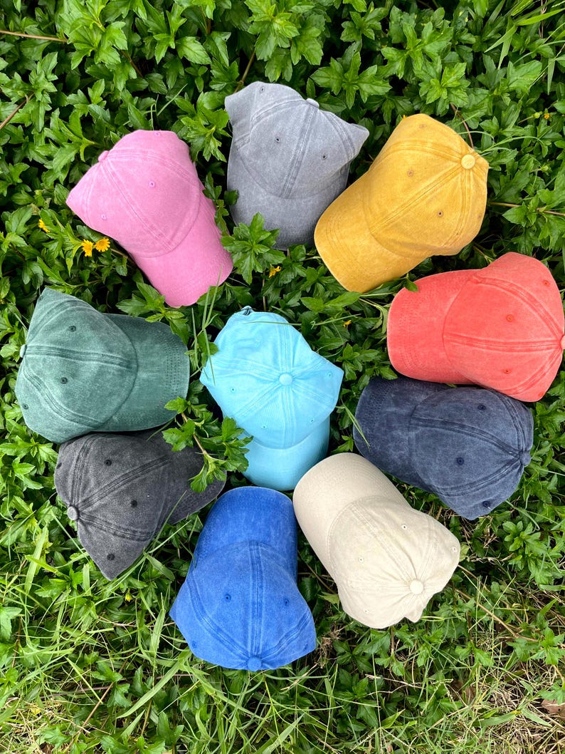 May include: Ten different colored baseball caps arranged in a circle on a bed of green grass. The caps are made of a soft, textured fabric and come in a variety of colors, including blue, green, red, pink, yellow, and gray.