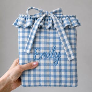 May include: A quilted pouch featuring a blue and white gingham pattern with a ruffled top edge and a matching fabric tie. The name Emily is embroidered in blue script on the front of the pouch, which has vertical stitching throughout the design.