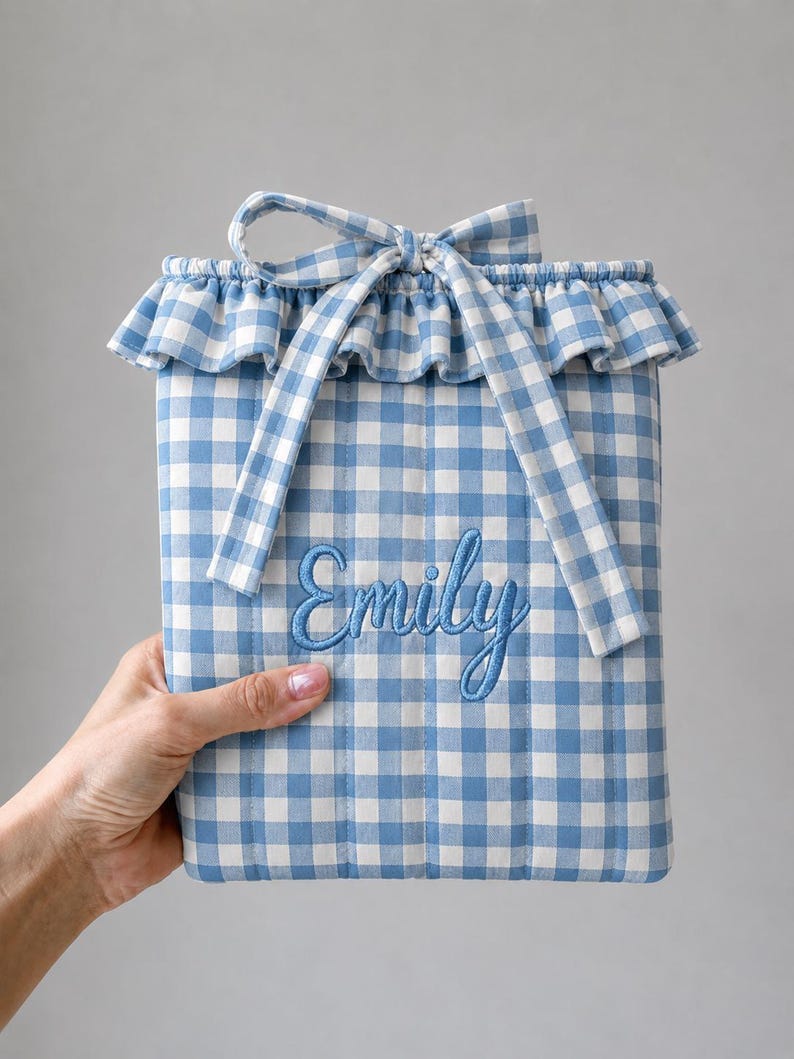 May include: A quilted pouch featuring a blue and white gingham pattern with a ruffled top edge and a matching fabric tie. The name Emily is embroidered in blue script on the front of the pouch, which has vertical stitching throughout the design.