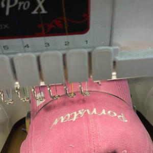 K&ouml;nnte beinhalten: Eine rosafarbene Baseballkappe mit dem Wort "bonjour" in wei&szlig; auf der Vorderseite gestickt. Die Kappe wird auf einer Stickmaschine gestickt.