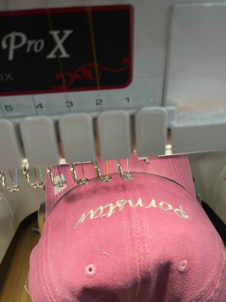 K&ouml;nnte beinhalten: Eine rosafarbene Baseballkappe mit dem Wort "bonjour" in wei&szlig; auf der Vorderseite gestickt. Die Kappe wird auf einer Stickmaschine gestickt.