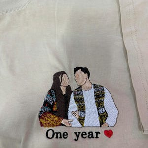 May include: A beige t-shirt featuring an embroidered design of two faceless figures standing side by side. The figure on the left wears a dark patterned garment, while the figure on the right wears a white shirt and a patterned vest. The text One year and a red heart are embroidered below.