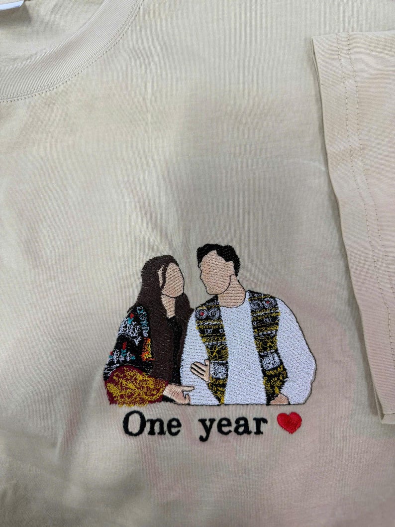 May include: A beige t-shirt featuring an embroidered design of two faceless figures standing side by side. The figure on the left wears a dark patterned garment, while the figure on the right wears a white shirt and a patterned vest. The text One year and a red heart are embroidered below.
