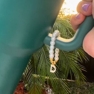 Può includere: Un braccialetto di perle bianche con un ciondolo dorato a forma della lettera "D". Il braccialetto è appeso al manico di una borraccia Stanley verde.