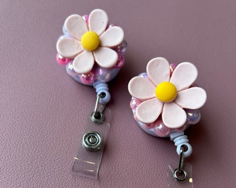 May include: Two daisy-shaped badge reels. Each reel has a light pink flower with a yellow centre, surrounded by iridescent beads. The reels have a clear plastic strap and a metal clip for attachment.