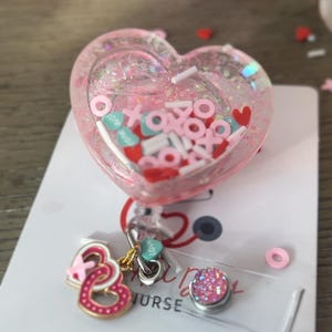 Puede incluir: Portagafete en forma de corazón rosa con purpurina y confeti de colores en el interior. El portagafete tiene un clip retráctil y un amuleto de doble corazón rosa y dorado. El texto "NURSE" es visible en el portagafete.