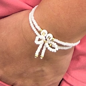 May include: White beaded bracelet with a gold 'RN' charm and a white bow accent.