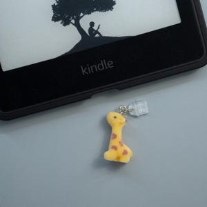 May include: A yellow giraffe charm with brown spots hangs near a Kindle e-reader. The e-reader is black and displays a silhouette of a tree and a person reading. The word "kindle" is printed on the device.