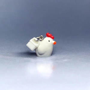 May include: A white ceramic chicken figurine with a red comb and orange beak. A small white bucket hangs from a silver-colored ring attached to the chicken's back. The figurine is set against a soft, gray background.