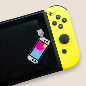 May include: A clear acrylic charm in the shape of a handheld gaming device, featuring a colorful abstract design of pink, blue, and yellow. The charm is attached to a keychain and is placed on a black screen and a yellow game controller.