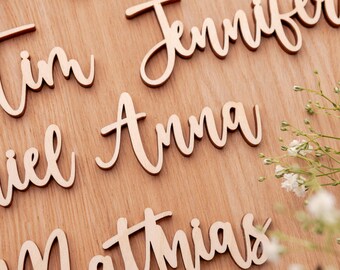 Puede incluir: Nombres de madera cortados con láser en fuente cursiva, esparcidos sobre una superficie de madera marrón claro. Los nombres incluyen "Jennifer", "Anna", "Matthias" y otros. Una ramita de flores blancas está en la esquina.