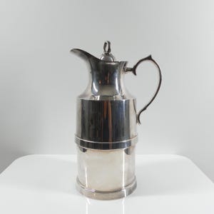 Op de afbeelding: Een zilverkleurige kan met een gebogen tuit, een handvat en een deksel met een kleine knop. De kan heeft een cilindrisch lichaam met een horizontale band in het midden. De kan staat op een wit oppervlak.