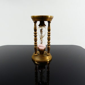 Op de afbeelding: Een zandloper van messing met een roze zandteller. De zandloper staat op een zwart oppervlak.