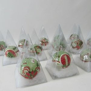 Könnte beinhalten: Eine Sammlung von 12 grünen und roten Weihnachtskugeln mit aufwendigen Designs, die in durchsichtigen dreieckigen Boxen mit weißer Baumwollfüllung präsentiert werden.