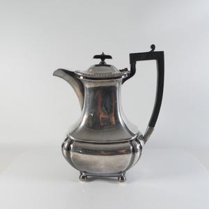 Op de afbeelding: Een gepolijste zilveren theepot met een gebogen tuit, een koepelvormig deksel met een kleine knop en een donker, hoekig handvat. Het lichaam heeft een afgeronde, gegroefde basis die rust op vier kleine, bolvormige voetjes.