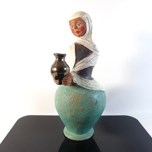 Könnte beinhalten: Eine Keramikskulptur einer Figur mit weißer Kopfbedeckung und dunklem Oberteil, die eine kleine, glänzend schwarze Vase hält. Die Figur hat einen strukturierten, türkisfarbenen Sockel mit abgerundeter Form. Das Gesicht hat blaue Augen.
