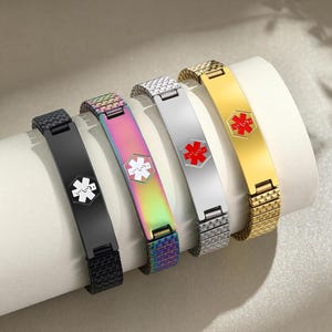 May include: Five medical alert bracelets in black, iridescent, silver, and gold. Each bracelet features a medical symbol in white or red. The bracelets are displayed on a white cylindrical surface.