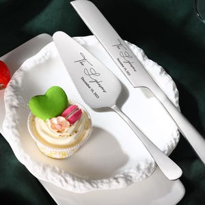以下が含まれることがあります： 刻印されたテキストが入った銀色のケーキナイフとサーバーのセット。装飾的な縁が付いた白いプレートの上に置かれています。緑のハート型の飾りと赤いトマトが付いた小さなカップケーキもあります。結婚式用です。