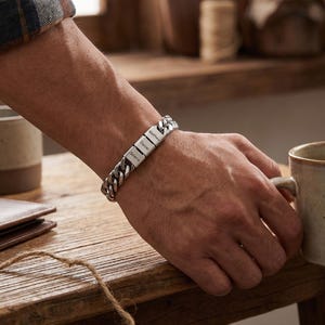 Puede incluir: Una pulsera de cadena de eslabones plateada con tres cuentas metálicas rectangulares, cada una grabada con un nombre: Selena, Daniel y Matthew. La pulsera tiene un acabado metálico pulido y un cierre seguro para llevar en la muñeca.