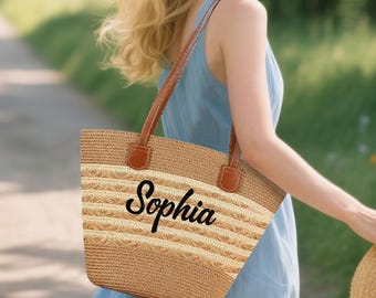May include: A woven straw tote bag with brown leather handles and the name "Sophia" in black script. The bag features horizontal stripes in natural and light beige tones. The bag is being carried by a person wearing a blue dress.