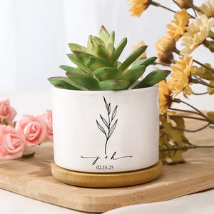 May include: A white ceramic planter with a green succulent plant. The planter has a black botanical design and the inscription "j + h 02.15.25". The planter sits on a round, light brown wooden coaster.
