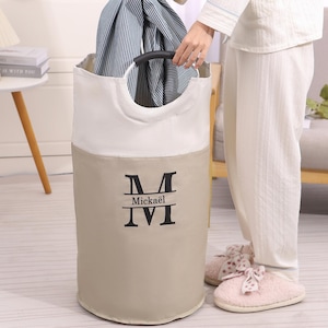 May include: A beige and white laundry hamper with black handles and the name "Mickaël" printed on the front. The hamper is cylindrical and contains blue and white striped clothing.