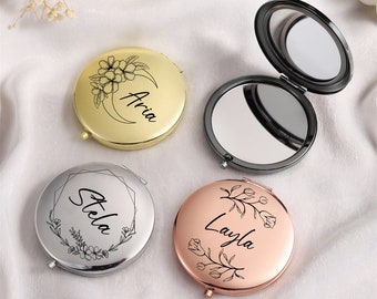 Può includere: Quattro specchietti compatti personalizzati in oro, argento e oro rosa. Ogni specchietto presenta un nome e un design floreale. Uno specchietto è aperto, rivelando la superficie riflettente.