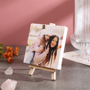 Puede incluir: Un marco de fotos blanco con una foto de una madre y una hija sonriendo. El marco está en un pequeño caballete de madera.