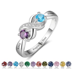 May include: A silver infinity ring with two gemstones, one purple and one blue. The ring is engraved with the words "Anna" and "Susan" and the text "Engraving Here".