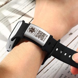 May include: A black medical ID bracelet with a silver plate featuring a medical symbol, QR code, and the text "HEALTH INFO SCAN ME." The bracelet is worn on a wrist.