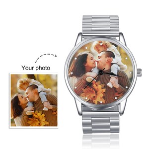 May include: A silver wristwatch with a round face featuring a photo of a family of three. The photo shows a couple holding their child, with the child's arms around the father's neck. The watch face has a silver bezel with small rhinestones around the edge.