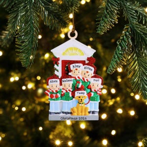 Peut inclure: Une décoration de sapin de Noël en forme de façade de maison avec cinq personnes et un chien portant des bonnets et écharpes rouges et blancs. Les prénoms Alola, Melissa, Alex, Ellen, Paul et Teddy sont inscrits. La base indique Christmas 2026.