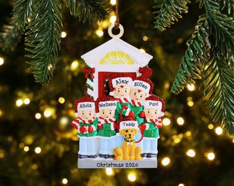 May include: A Christmas tree ornament shaped like a house front with five people and a dog wearing red and white hats and scarves. The names Alola, Melissa, Alex, Ellen, Paul, and Teddy are printed on the hats. The base reads Christmas 2026.