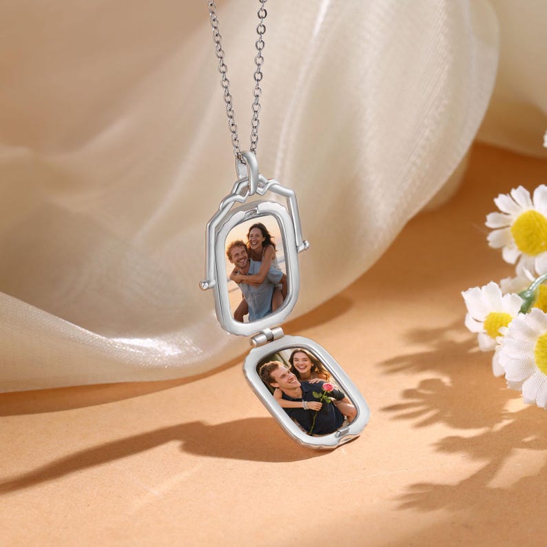 May include: A silver locket necklace with two rectangular compartments, each displaying a photograph. The locket is open, revealing the images of two couples. The necklace hangs against a soft, neutral background, with a few daisies in the corner.