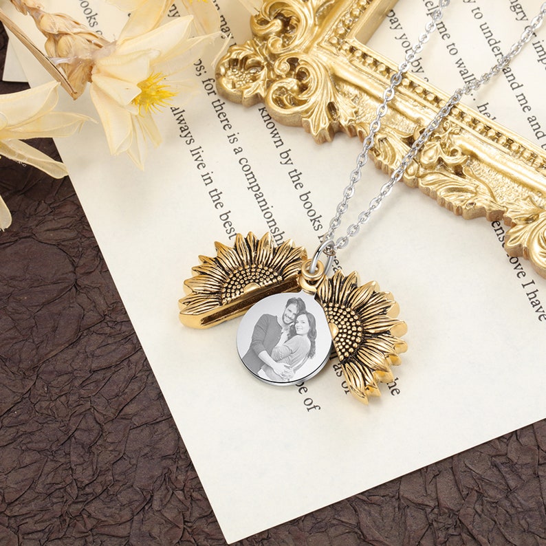 Pu&ograve; includere: Una collana con ciondolo a forma di girasole dorato con una catena d'argento. Il ciondolo si apre per rivelare una foto in bianco e nero di una coppia. I petali del girasole sono dettagliati.
