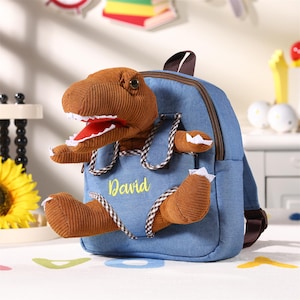 May include: A blue denim backpack featuring a brown corduroy dinosaur design. The dinosaur's head and limbs extend from the front pocket. The name "David" is embroidered in yellow on the pocket.