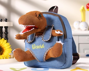 May include: A blue denim backpack with a brown corduroy dinosaur emerging from the front pocket. The dinosaur has a red mouth and white teeth. The name "David" is embroidered in yellow on the pocket.