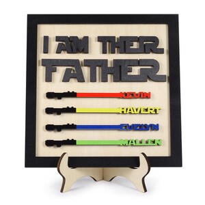 Puede incluir: Un letrero de madera con el texto "I AM THEIR FATHER" en letras negras. El letrero tiene cuatro sables de luz con los nombres "Kevin", "Havert", "Evelyn" y "Mallen" escritos en ellos en diferentes colores.