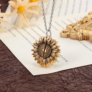 Pu&ograve; includere: Collana con pendente a forma di girasole dorato e catena argentata. Il pendente a forma di girasole presenta petali strutturati e un motivo centrale a seme. La collana &egrave; esposta su una superficie color crema.